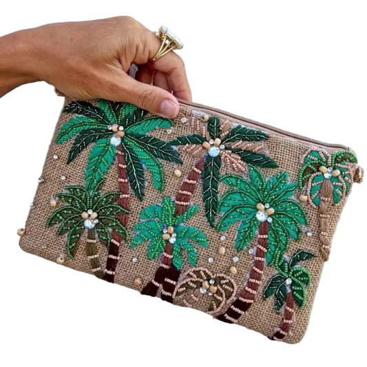 A hand holds a small zip pouch decorated with embroidered palm trees and small pearl-like beads on a tan woven background, against a plain light wall.