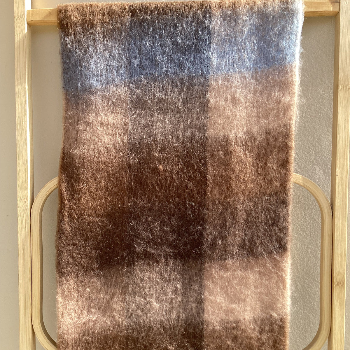 A close-up of a fluffy, soft-textured plaid scarf or throw in muted brown, beige, and blue-gray tones, draped over a light wood rack.