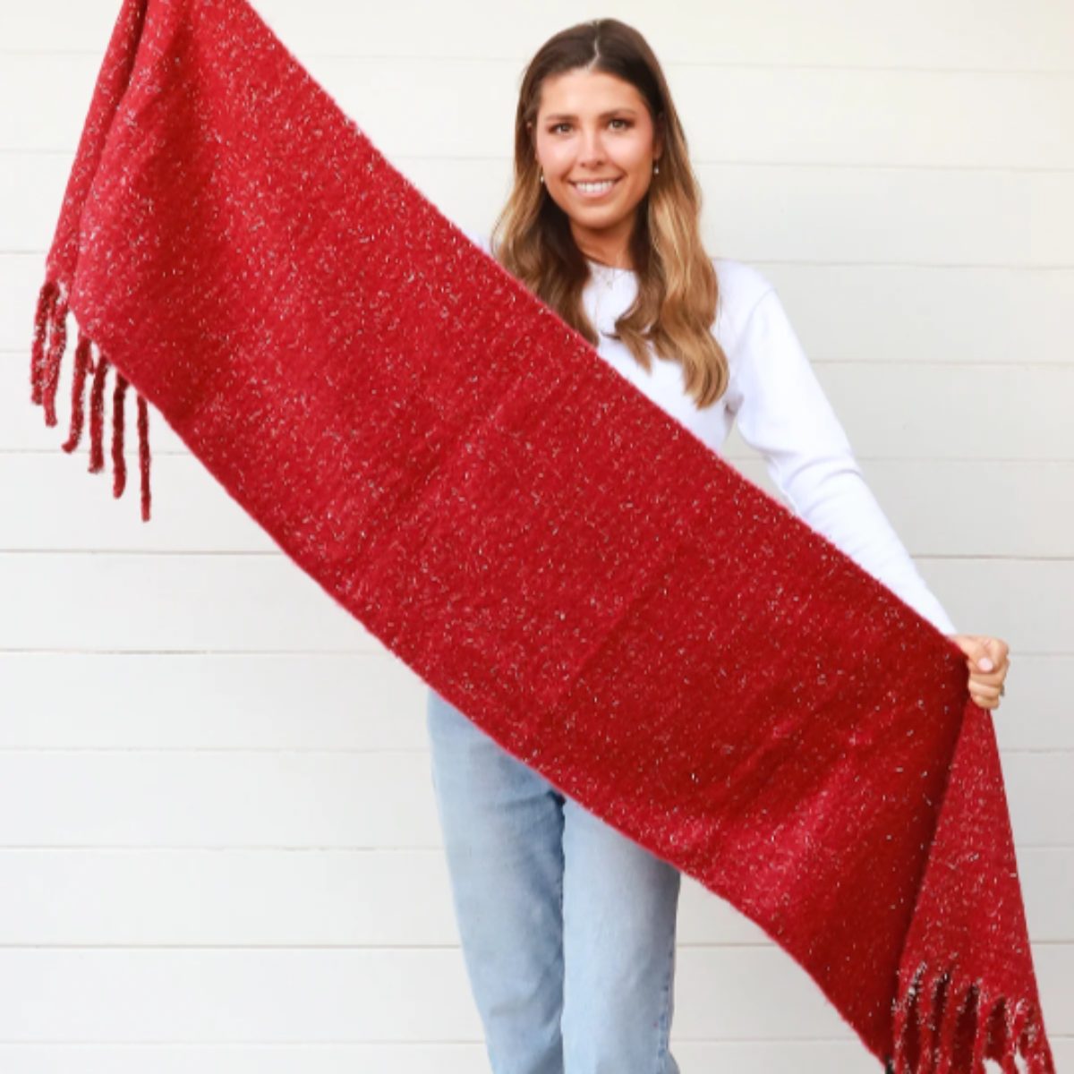 Woman holding a large red scarf with tassels against a white wall.
