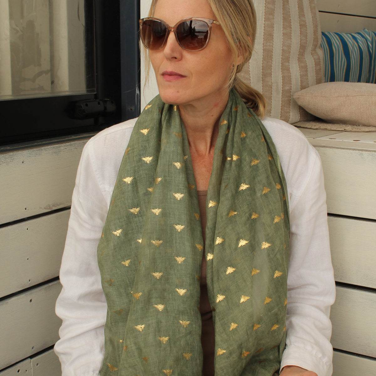 A woman wearing a sage green scarf with small gold metallic bee motifs, styled with a white long-sleeve shirt and sunglasses, sitting on a white wooden porch.