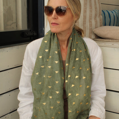 A woman wearing a sage green scarf with small gold metallic bee motifs, styled with a white long-sleeve shirt and sunglasses, sitting on a white wooden porch.