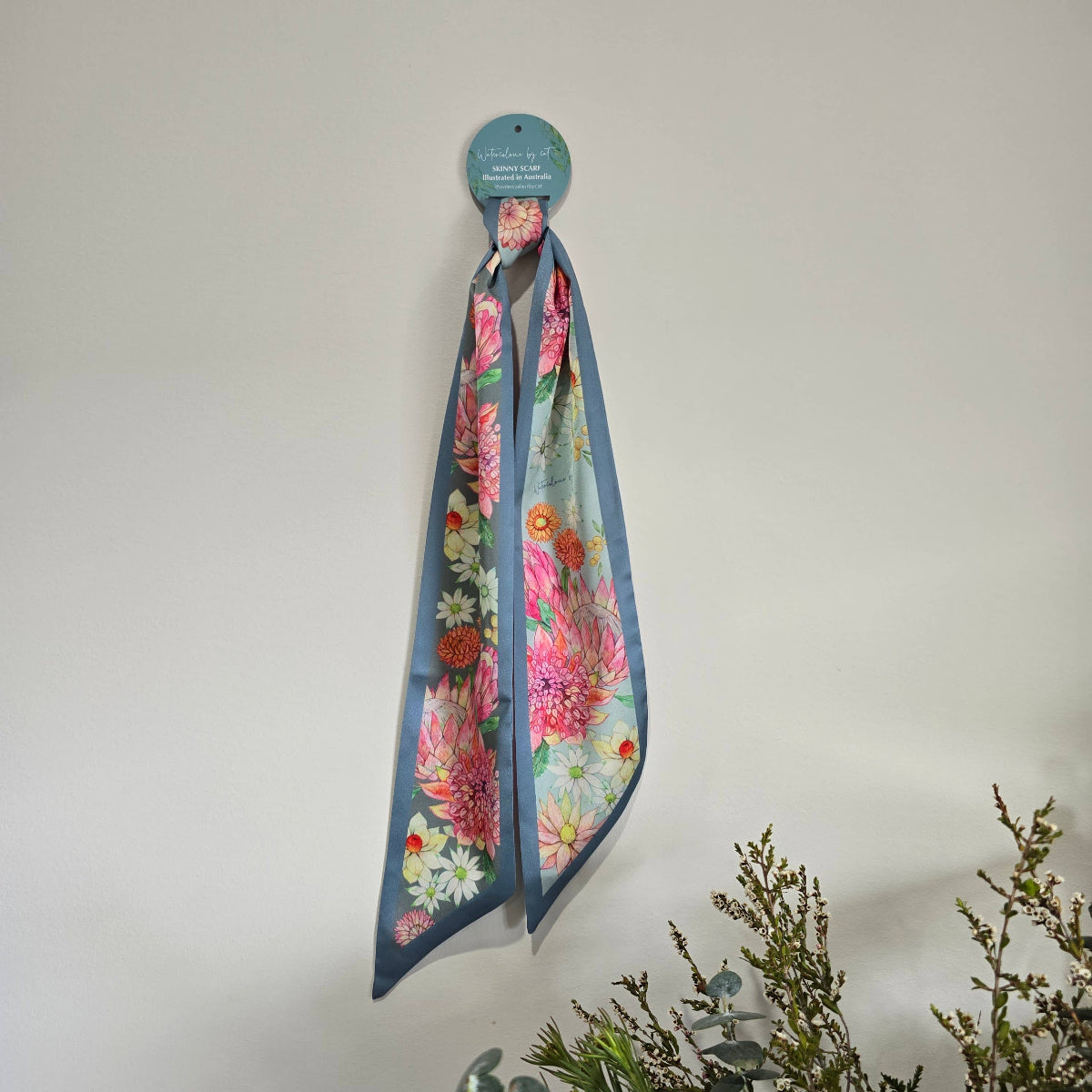 A floral silky scarf with a dusty blue border hangs from a circular teal display card against a plain off-white wall, with decorative greenery in the lower right corner.