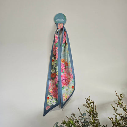 A floral silky scarf with a dusty blue border hangs from a circular teal display card against a plain off-white wall, with decorative greenery in the lower right corner.