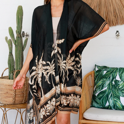 Woman wearing a black floral kimono in a tropical-themed room with a cactus and woven chair.