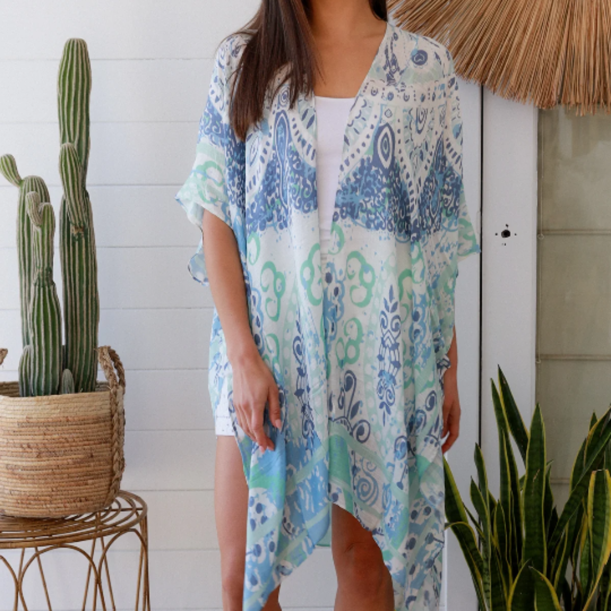 Woman wearing a blue and green kimono standing in a room with plants and a decorative wall piece.