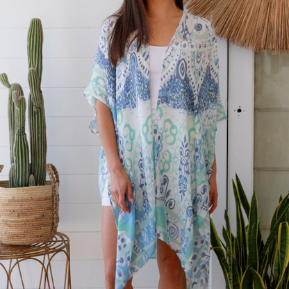Woman wearing a blue and green kimono standing in a room with plants and a decorative wall piece.