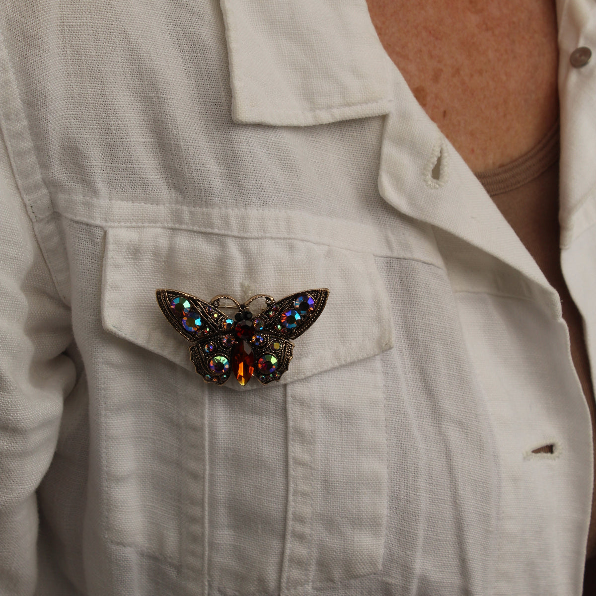 A close-up of a person wearing a white linen shirt, featuring a detailed butterfly brooch pinned to the chest pocket. The brooch is crafted with dark metal, iridescent blue and green rhinestones, and a central amber-orange stone.