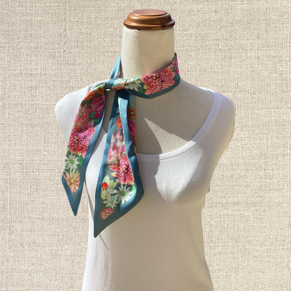 A floral silk neck scarf with a teal base and pink, red, and yellow flowers, styled on a cream-colored mannequin torso against a neutral textured beige background.