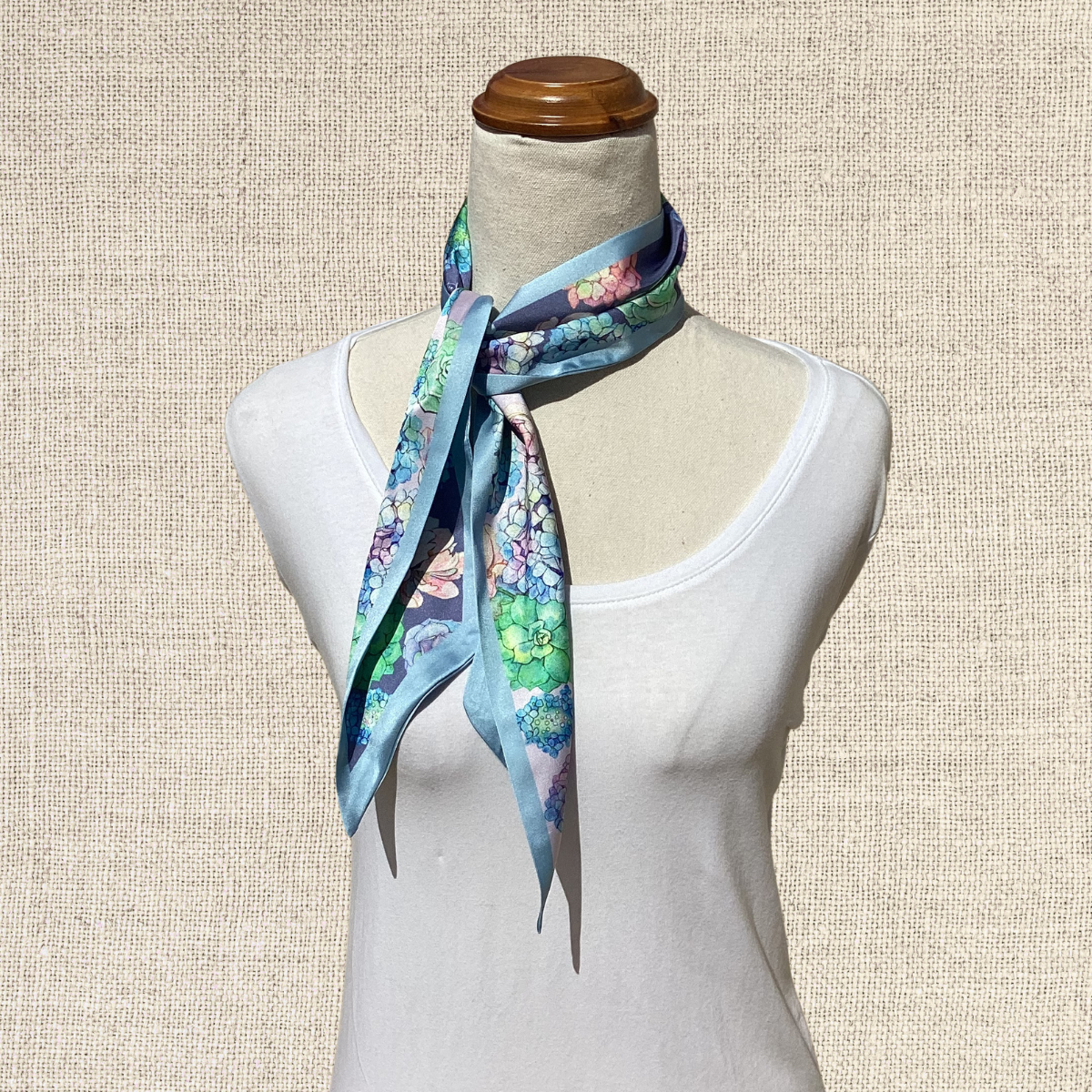 A light blue silk scarf with a colorful floral and butterfly print, styled on a cream-colored mannequin torso against a textured beige background.