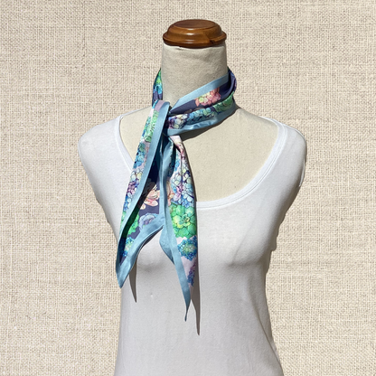 A light blue silk scarf with a colorful floral and butterfly print, styled on a cream-colored mannequin torso against a textured beige background.