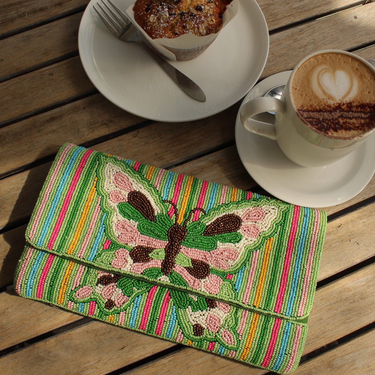 A colorful striped clutch bag with a large beaded butterfly design in green, pink, and brown, styled on a wooden table next to a cup of coffee and a muffin.