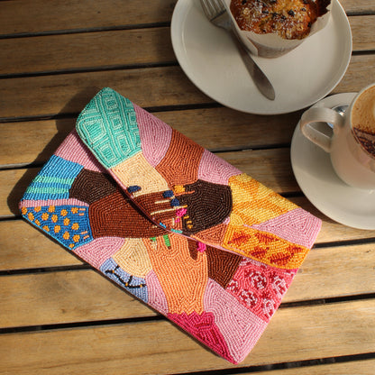 A colorful beaded clutch bag with holding hands print, styled on a wooden table alongside coffee cups and a muffin.