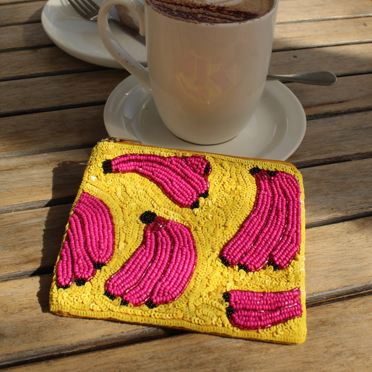 A yellow sequin purse with bright pink beaded banana motifs, placed on a wooden cafe table next to a cup of cappuccino and pastries.