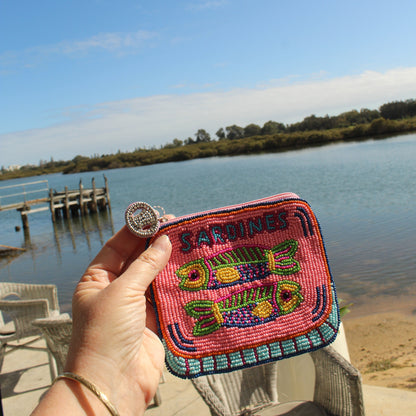 A hand holds a small embroidered coin purse featuring colorful fish designs and the word 'SARDINES' on a pink background, with a lake and dock in the background.