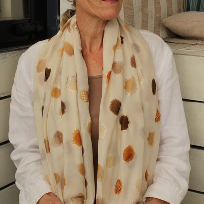 A woman wearing a cream-colored scarf with brown and gold abstract circular patterns, styled with a white shirt and sunglasses.