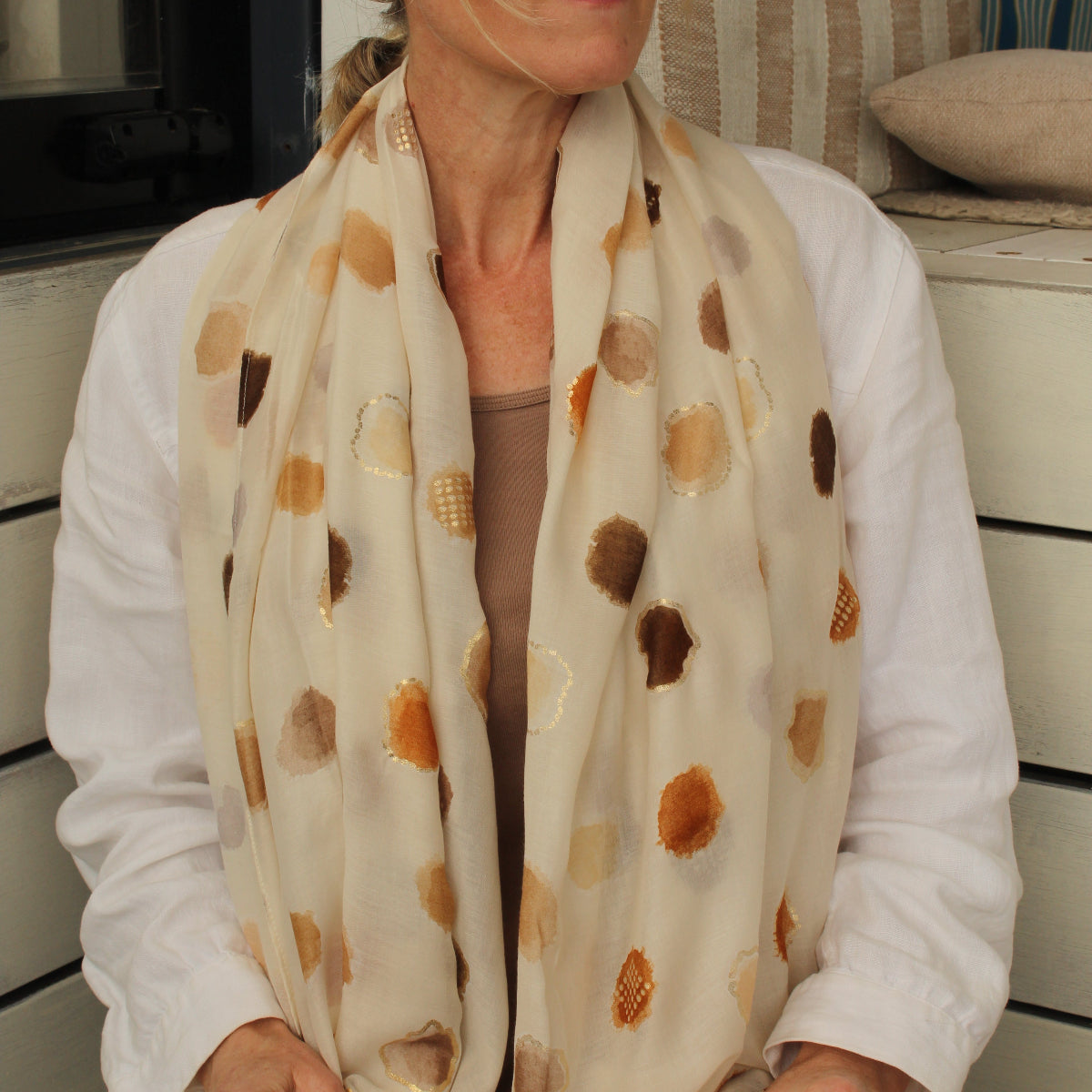 A woman wearing a cream-colored scarf with brown and gold abstract circular patterns, styled with a white shirt and sunglasses.