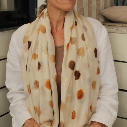 A woman wearing a cream-colored scarf with brown and gold abstract circular patterns, styled with a white shirt and sunglasses.