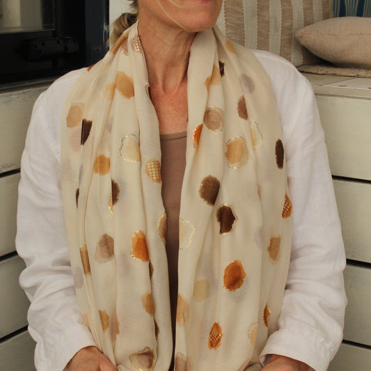 A woman wearing a cream-colored scarf with brown and gold abstract circular patterns, styled with a white shirt and sunglasses.