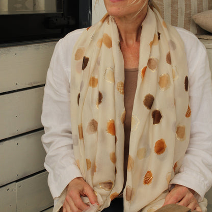 A woman wearing round sunglasses and a cream scarf with brown and orange polka dots, sitting in front of a white wooden wall.