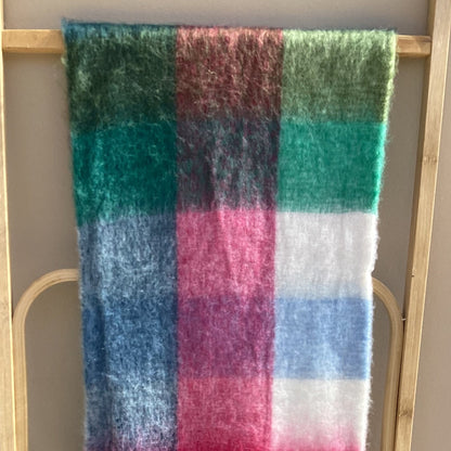 A plaid scarf with fringe, featuring stripes of green, blue, pink, white, and brown, draped over a light wood rack against a beige wall.