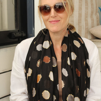A woman with blonde hair wearing round sunglasses, a white long-sleeve shirt, and a black scarf with abstract gold, brown, and beige circular patterns, posed in front of a striped cushion.