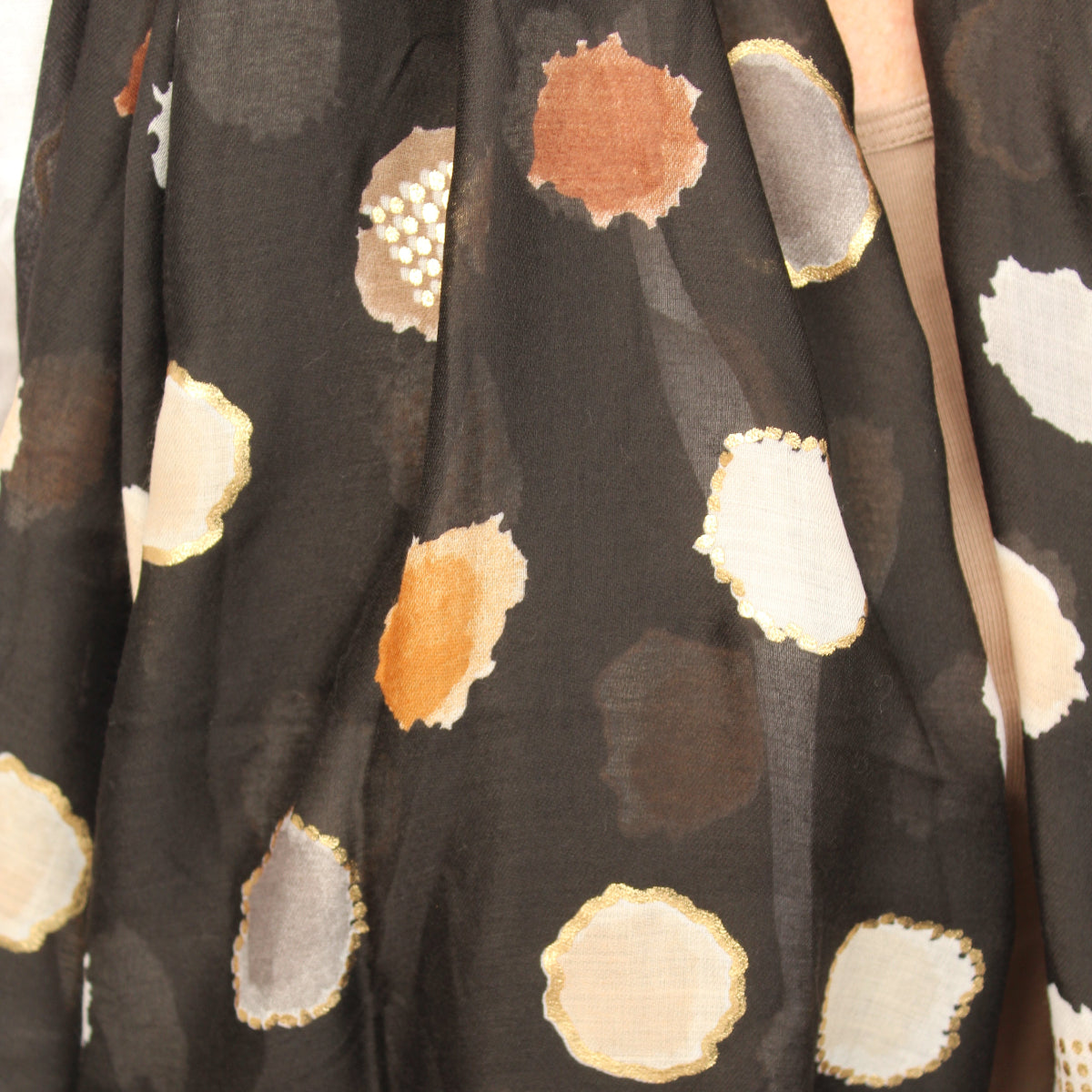 A close-up of a black scarf featuring a pattern of irregular watercolor-style polka dots in beige, white, brown, and gray.