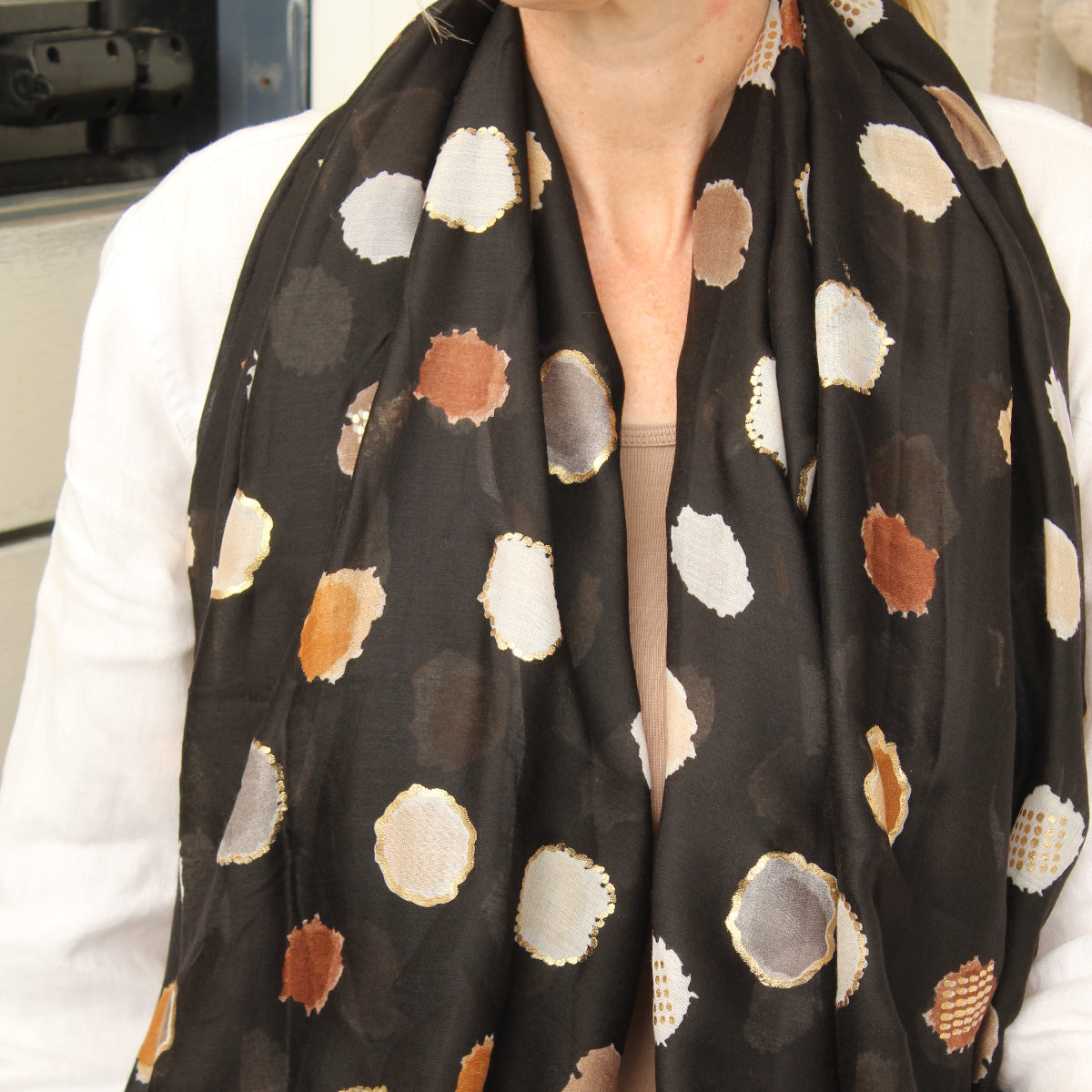 A black scarf with a pattern of irregular polka dots in brown, beige, and white, worn by a model in a white cardigan.