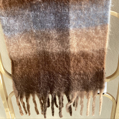 A fluffy plaid scarf in shades of brown, beige, and muted blue-gray, draped over a light wood hanger against a light wall.