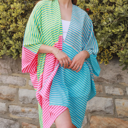 Woman wearing a colorful striped kimono-style garment against a stone wall and green bush background.