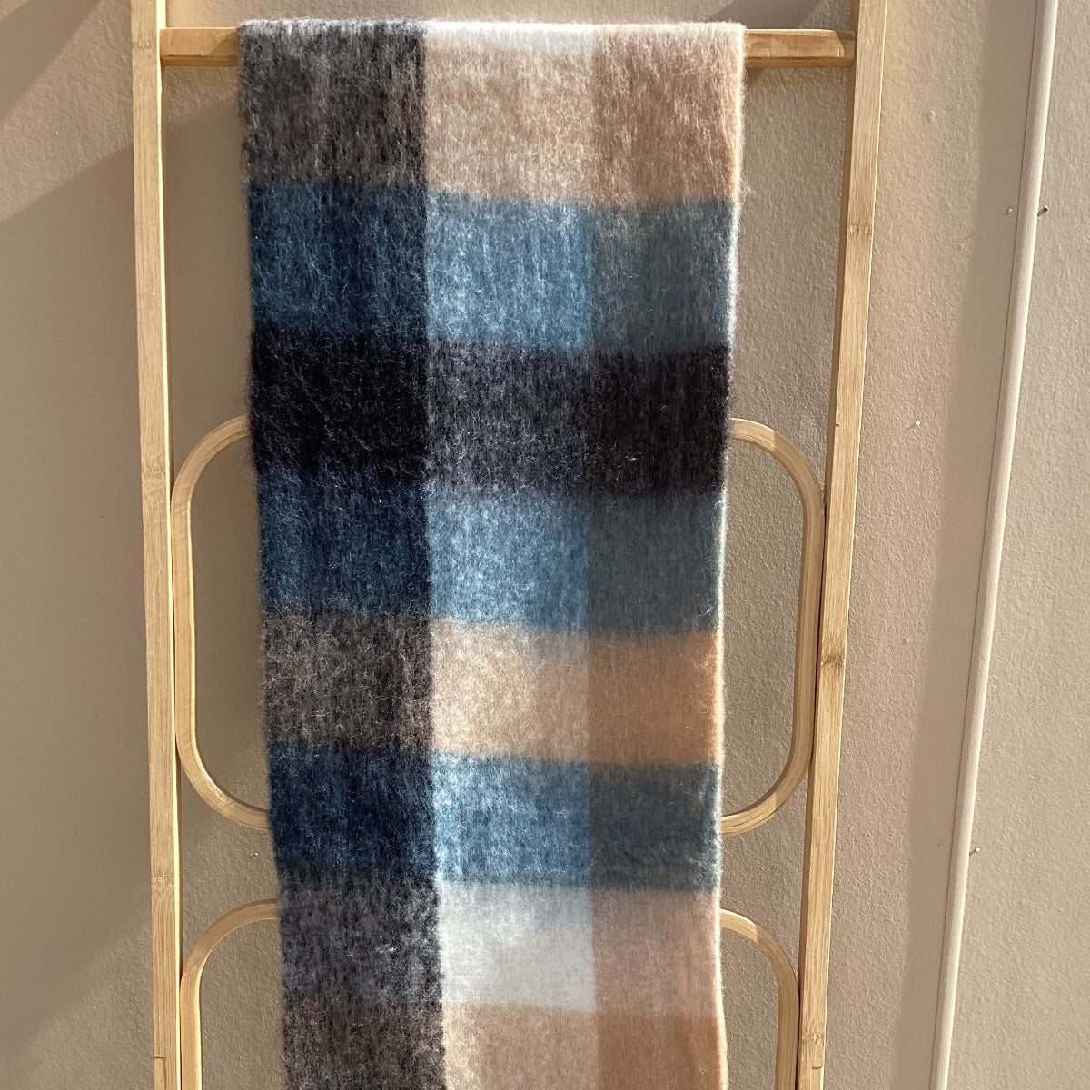 A soft woven scarf with a plaid pattern in shades of blue, beige, brown, and black, draped over a light wood coat rack against a beige wall.