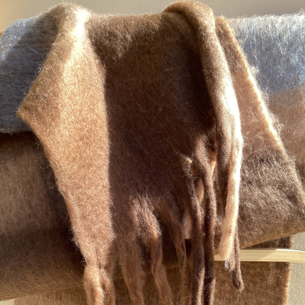 A close-up of a soft, fluffy scarf or wrap in warm taupe and brown tones, draped with fringed edges and a plaid pattern partially visible in the background.