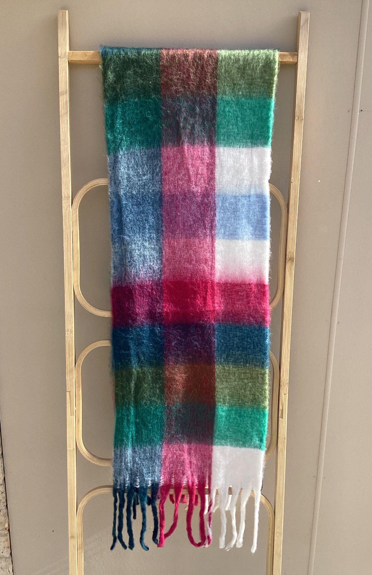 A multicolor plaid scarf with fringe, draped over a light wood ladder against a beige wall.