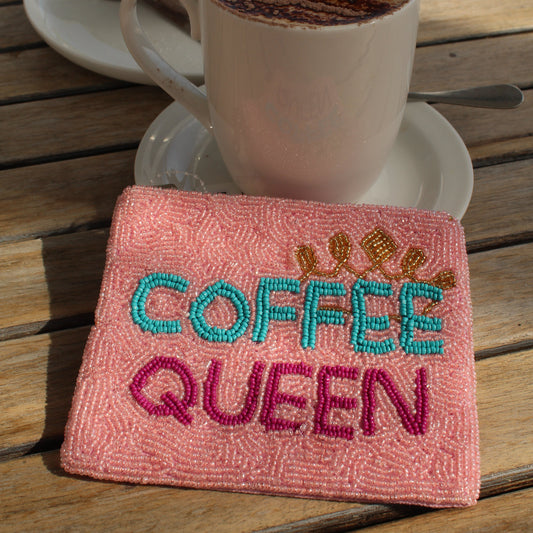 A pink beaded zip purse embroidered with 'COFFEE QUEEN' in turquoise and magenta, placed on a wooden table next to a white coffee mug with latte art and a muffin on a plate.