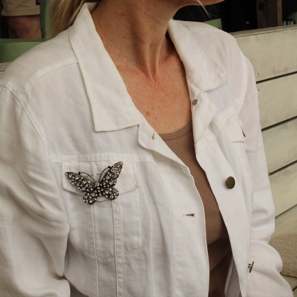 A woman in profile wearing a white denim jacket with a decorative butterfly brooch on the chest pocket, set against an outdoor patio background.