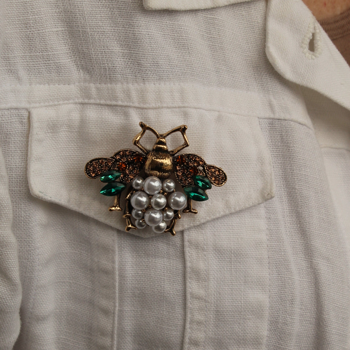A close-up of a vintage-style bee brooch pinned to the chest pocket of a white linen shirt. The brooch features a gold-toned body, dark brown wings, green enamel accents, and a cluster of white faux pearls.