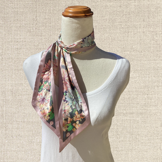 A floral silk scarf with a dusty pink base and multicolor flower print, styled on a white mannequin torso against a textured beige background.