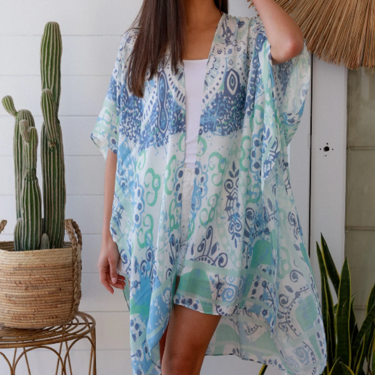 Woman wearing a blue and green patterned kimono standing in a room with plants and a decorative wall piece.