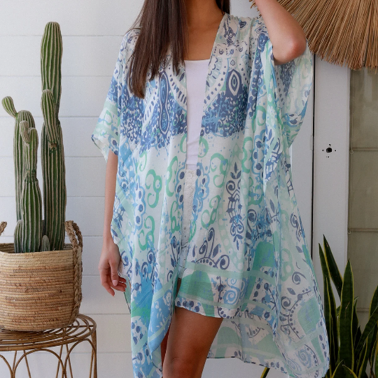 Woman wearing a blue and green patterned kimono standing in a room with plants and a decorative wall piece.