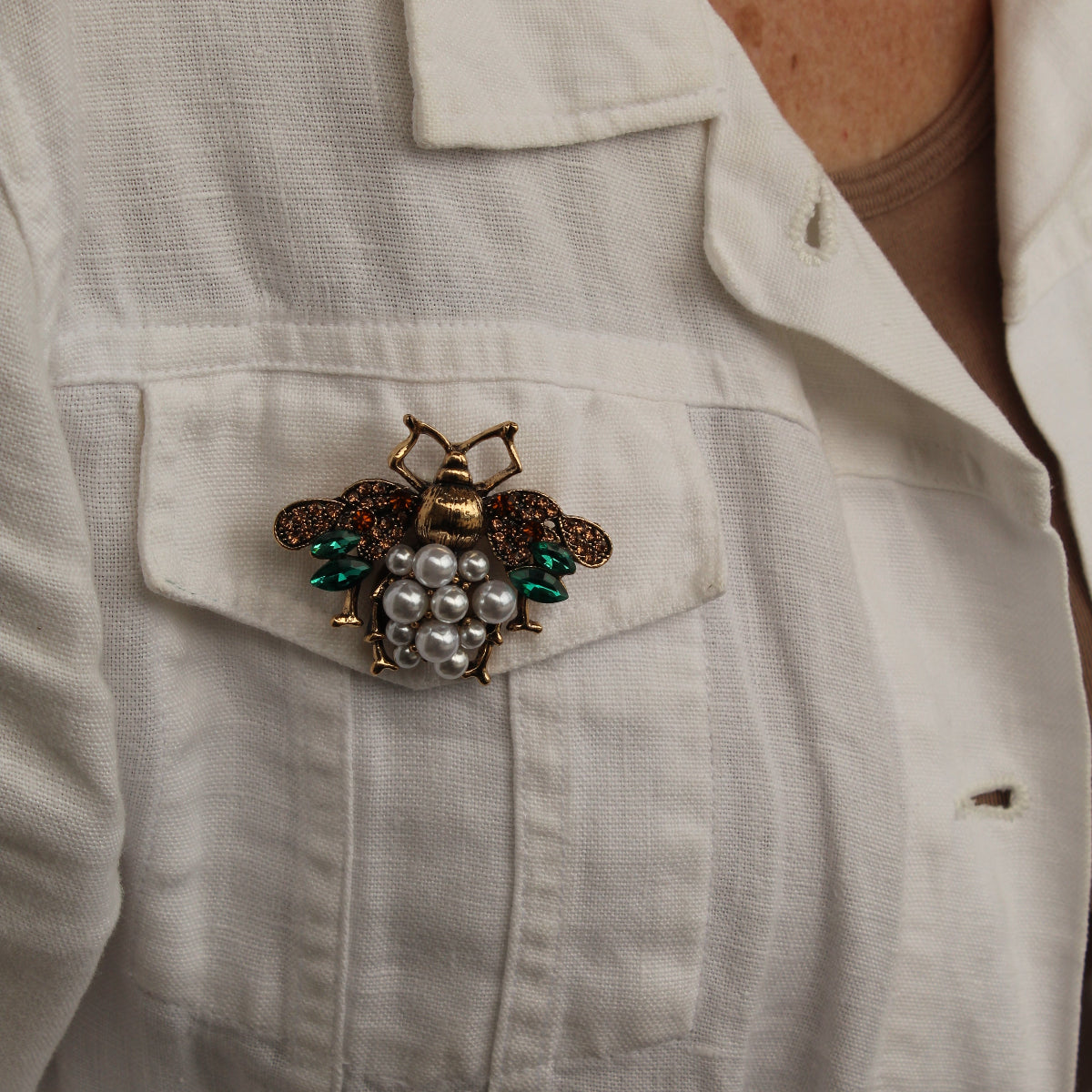 a close up of a woman wearing a white jacket and a bee brooch feautring pearls and green wings