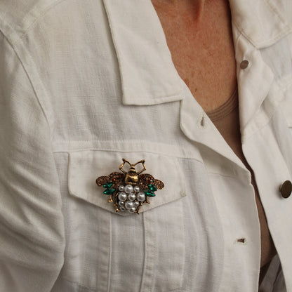 A close-up of a woman wearing a white linen button-up shirt, featuring a decorative brooch shaped like a grapevine with white faux pearls and green leaves pinned to the chest pocket.