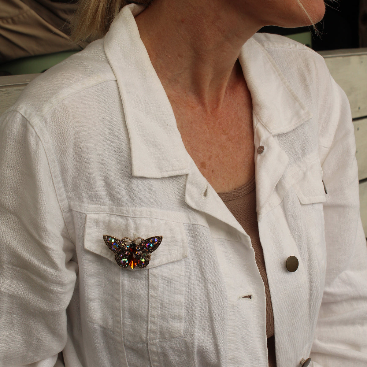 A close-up side profile of a woman wearing a white linen jacket with a colorful butterfly brooch pinned to the chest pocket.