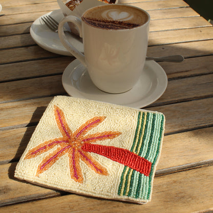 A close-up of a beaded zip purse on a wooden cafe table, featuring a red and orange palm tree design with green stripes. A white coffee cup on a saucer sits behind it on a wooden table with chairs in the background.