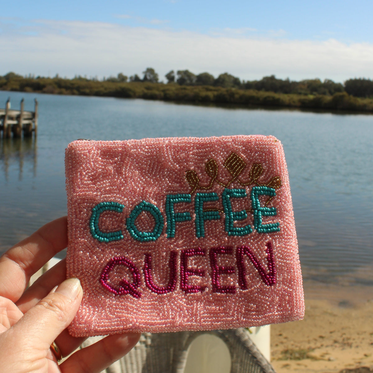 A hand holds a square pink embroidered purse with the words 'COFFEE QUEEN' in teal and magenta, with a small crown above the text, set against a background of a lake, dock, and patio chair.