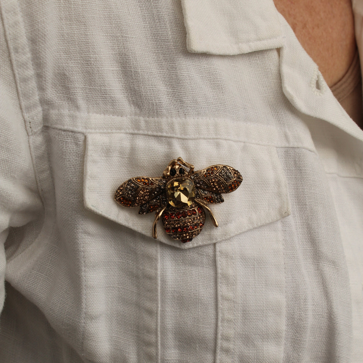 A close-up of a person wearing a white linen shirt, featuring a detailed bee brooch pinned to the chest pocket.