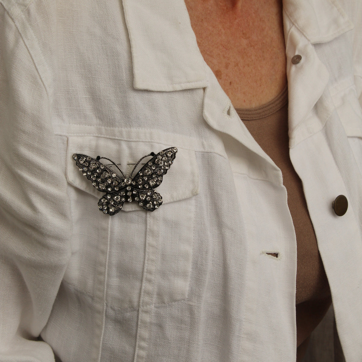 A close-up of a person wearing a white linen jacket over a beige top, featuring a black butterfly brooch embellished with small rhinestones on the left chest pocket.