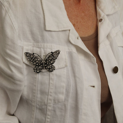 A close-up of a person wearing a white linen jacket over a beige top, featuring a black butterfly brooch embellished with small rhinestones on the left chest pocket.
