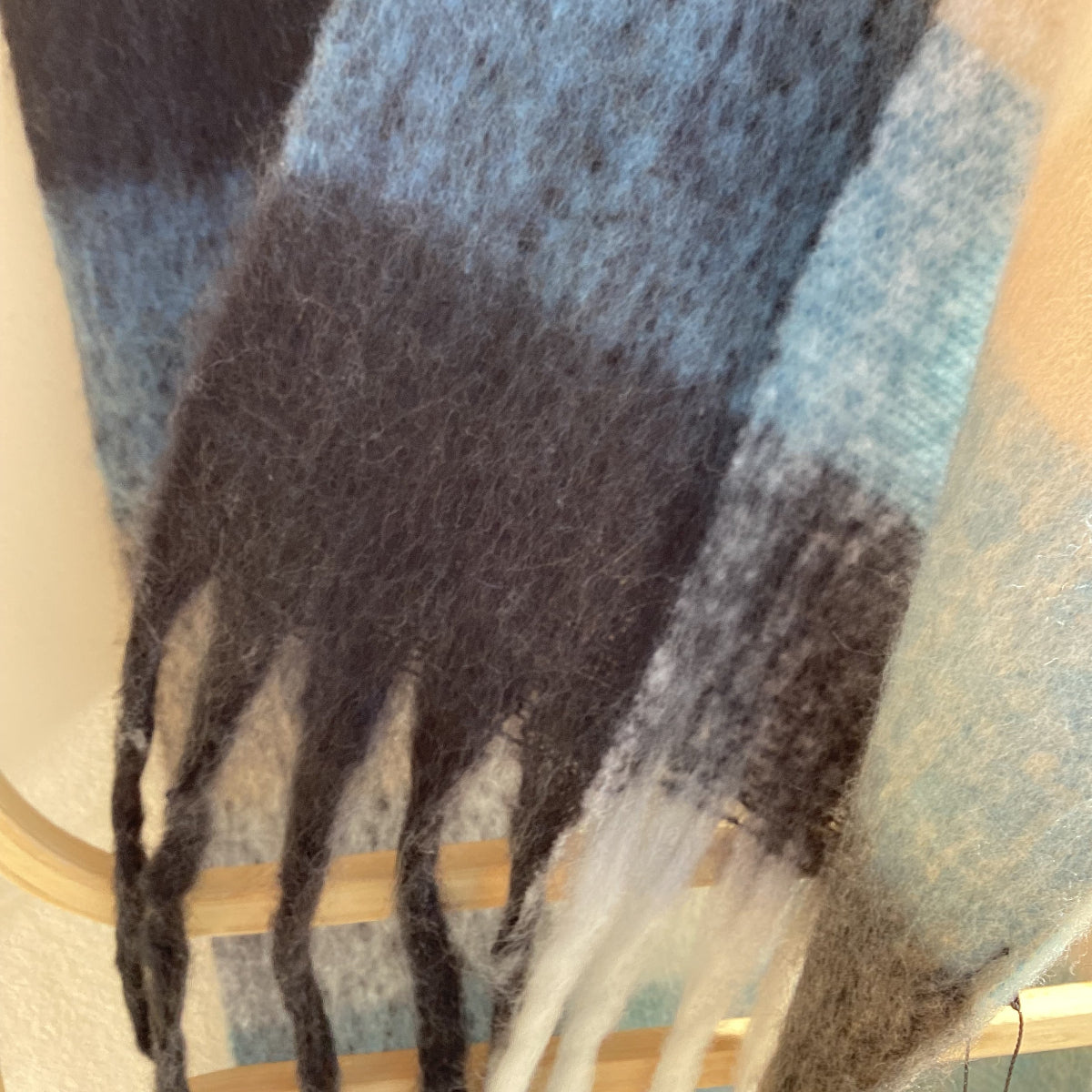 A close-up of a soft woven scarf or throw with a plaid pattern in light blue, charcoal gray, black, and cream, draped over a light wood hanger.