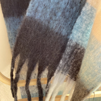 A close-up of a soft woven scarf or throw with a plaid pattern in light blue, charcoal gray, black, and cream, draped over a light wood hanger.