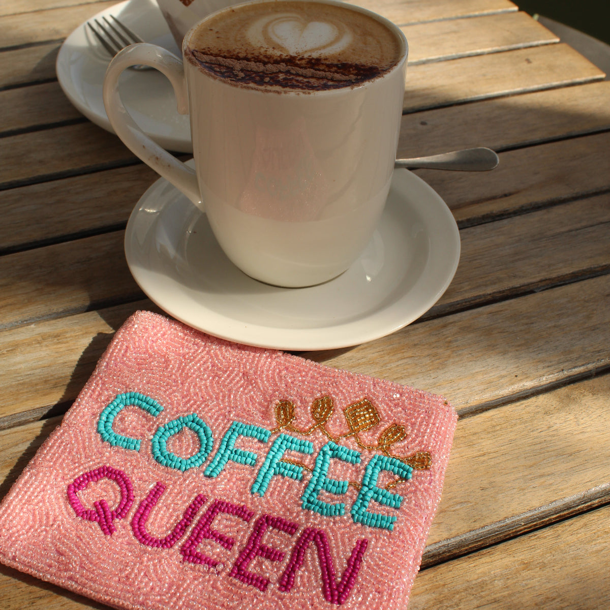 A pink embroidered zip purse with the words 'COFFEE QUEEN' in blue and magenta sits on a wooden outdoor table next to a white coffee cup on a saucer.