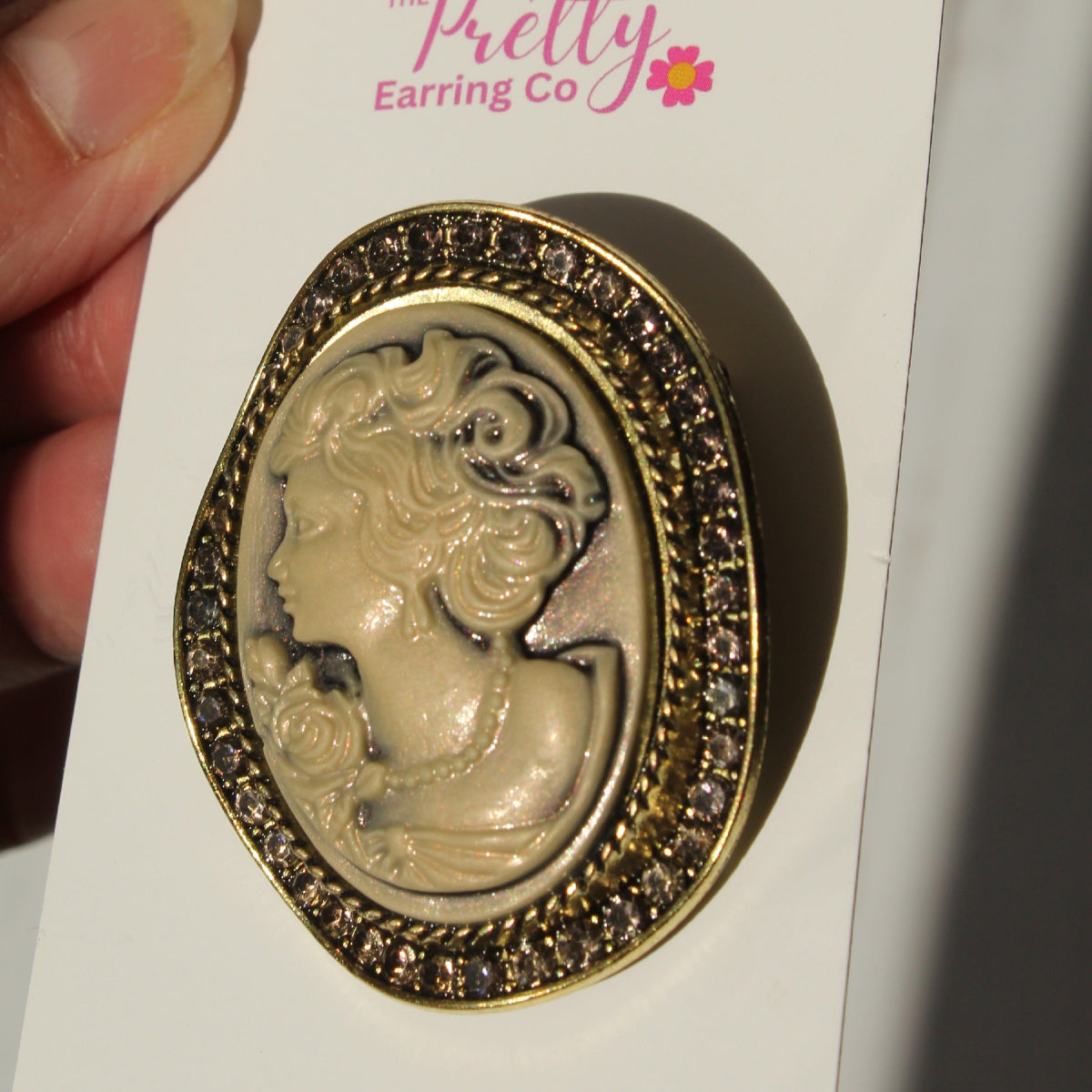 A vintage-style oval earring with a gold-tone frame and a beige embossed classical profile, mounted on a white display card held by a hand. The card is branded 'The Pretty Earring Co'.